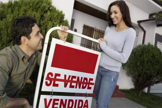 Dueños de casa felices enfrente de propiedad con letrero de vendida.