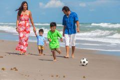 Familia hispana donde la madre, padre y dos hijos varones caminan en la playa siguiendo una pelota de futbol.