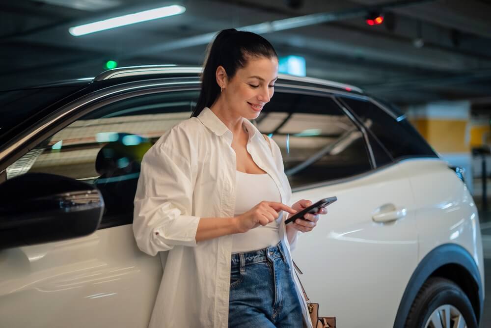 Mujer usando su teléfono junto a su auto en un estacionamiento, revisando información sobre registración en California.