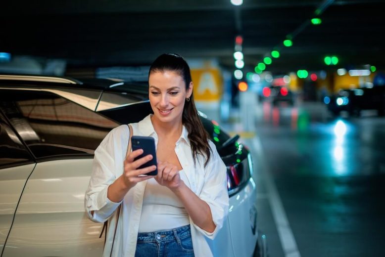 Mujer junto a su auto usando el celular en un estacionamiento, ideal para explicar el seguro de auto si vendes tu vehículo.