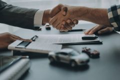 Acuerdo de venta de auto con apretón de manos sobre contrato, representando el seguro de auto y vehículo en proceso de venta.