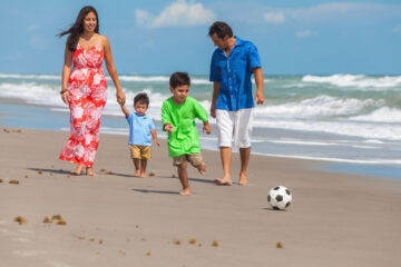 Familia hispana donde la madre, padre y dos hijos varones caminan en la playa siguiendo una pelota de futbol