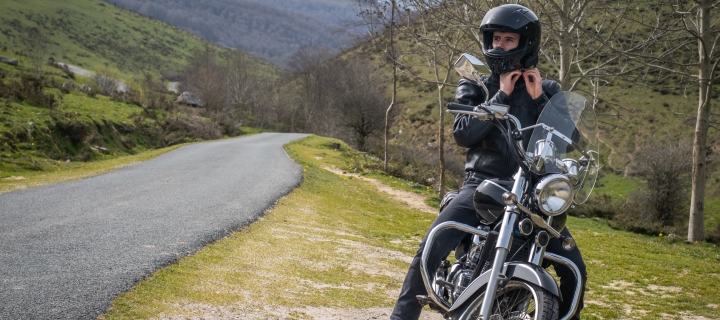 Joven motociclista poniéndose su casco negro junto a un camino de montaña y sentado en su motocicleta personalizada