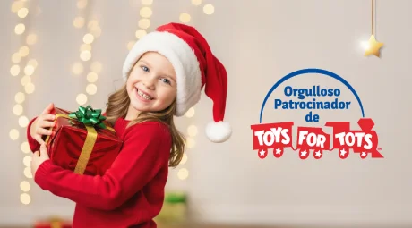 Una niña sonriente con un gorro de Santa y un suéter rojo sostiene un regalo envuelto frente a luces navideñas, junto al texto que dice “Orgulloso patrocinador de Toys for Tots”.
